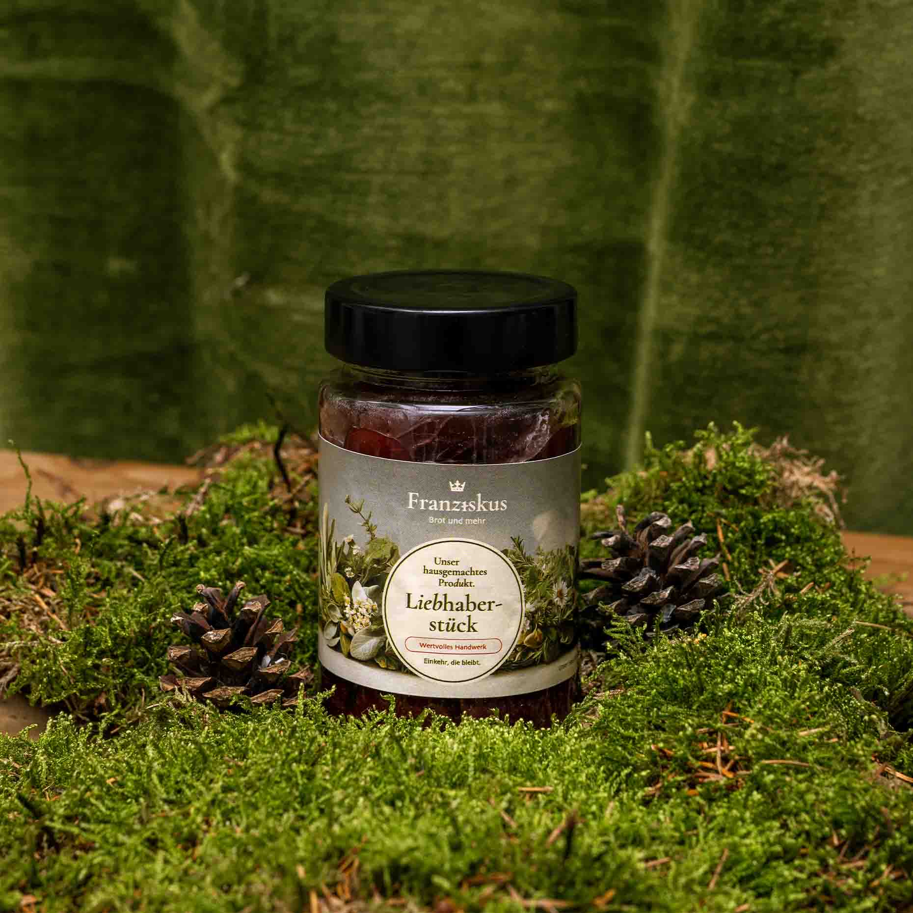 Ein Bild des hauseigenen Produkts: Rotweinzwetschgen – Liebhaberstück. Dekoriert auf Moos mit Kiefernzapfen und olivfarbenem Hintergrund.