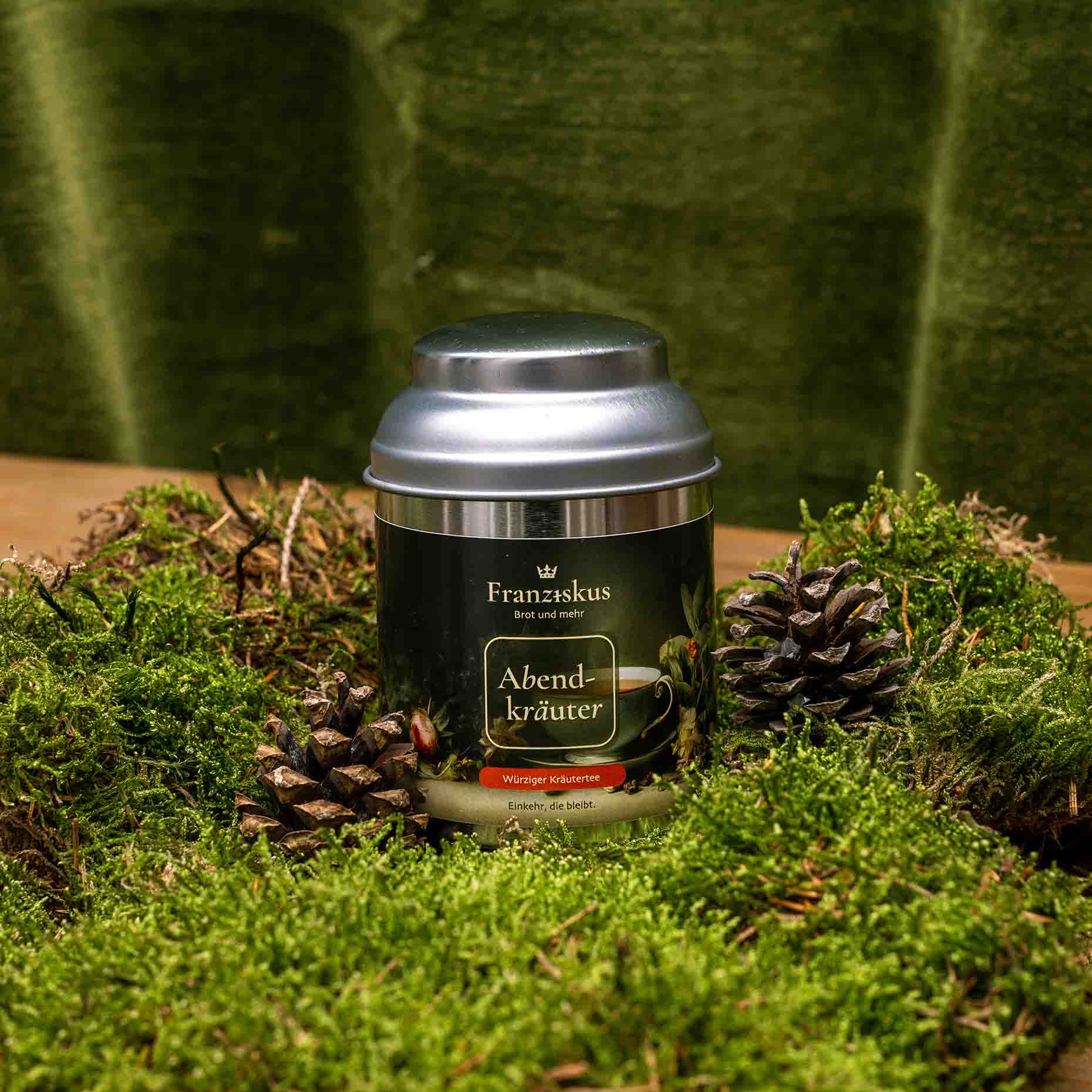 Ein Bild des Tees "Abendkräuter" des Haus Franziskus in der 50g Dose. Die Dose ähnelt in ihrer Form der Wallfahrtskirche in Freystadt. Ein würziger Kräutertee. Dekoriert auf Moos mit Kiefernzapfen und olivfarbenem Hintergrund.