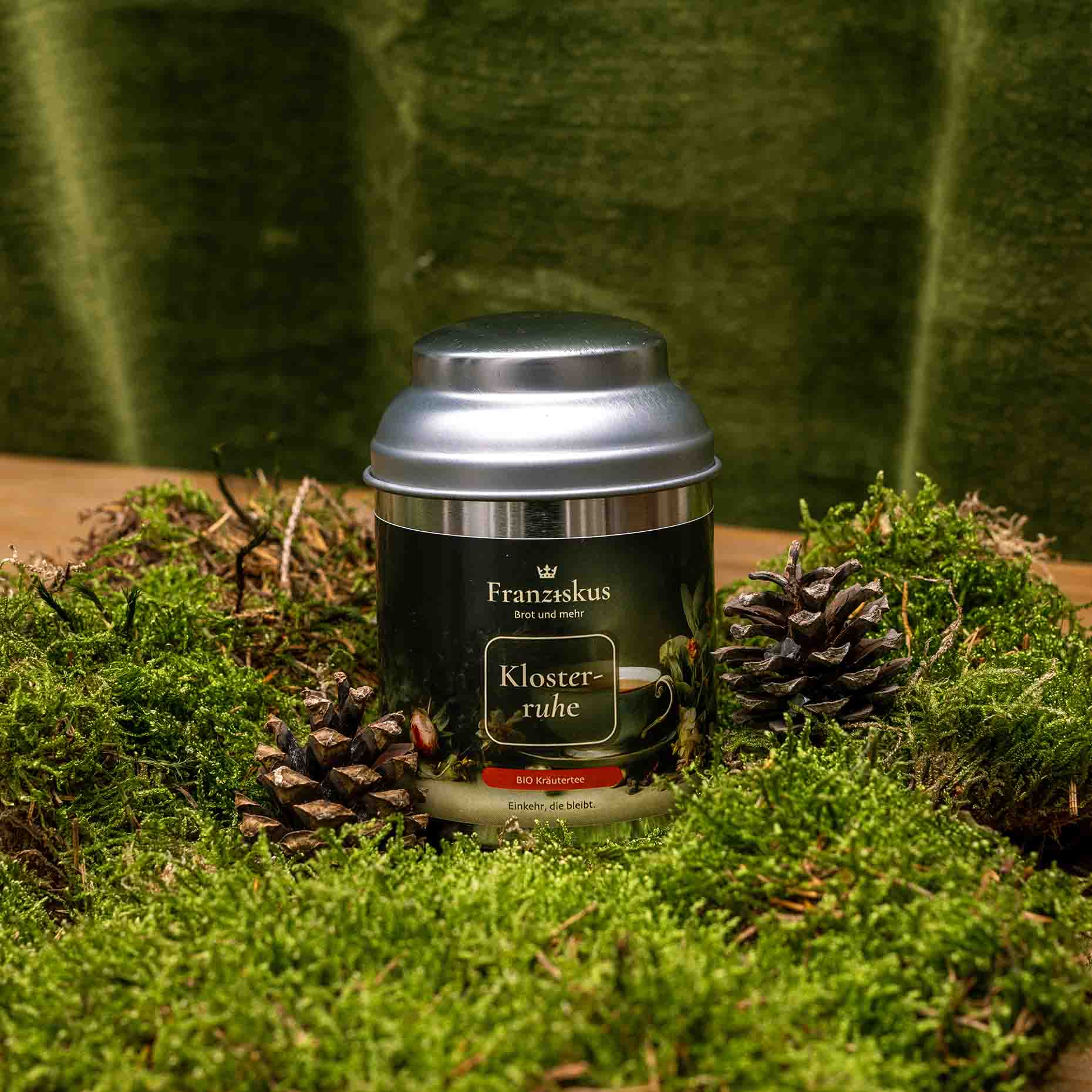 Ein Bild des eines Tees des Haus Franziskus: "Klosterruhe" in der 50g Dose. Die Dose ähnelt in ihrer Form der Wallfahrtskirche in Freystadt. Ein BIO Kräutertee. Dekoriert auf Moos mit Kiefernzapfen und olivfarbenem Hintergrund.