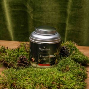 Ein Bild des Tees "Klosterruhe" des Haus Franziskus in der 125g Dose. Die Dose ähnelt in ihrer Form der Wallfahrtskirche in Freystadt. Ein BIO Kräutertee. Dekoriert auf Moos mit Kiefernzapfen und olivfarbenem Hintergrund.