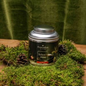 Ein Bild des Tees "Sieben Sinne" des Haus Franziskus in der 125g Dose. Die Dose ähnelt in ihrer Form der Wallfahrtskirche in Freystadt. Ein BIO Kräutertee. Dekoriert auf Moos mit Kiefernzapfen und olivfarbenem Hintergrund.